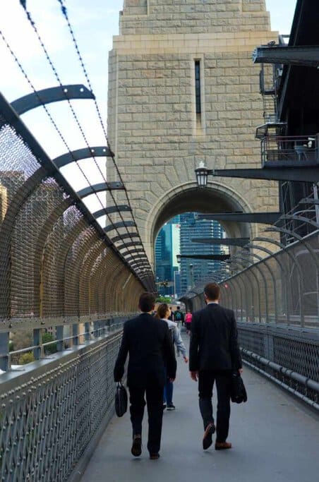 Commuters walking along the pedestrian pathway of the Sydney Harbour Bridge, bordered by safety fencing, with the city skyline visible through the arch, demonstrating the bridge's daily load in a depiction related to its weight-bearing capabilities.