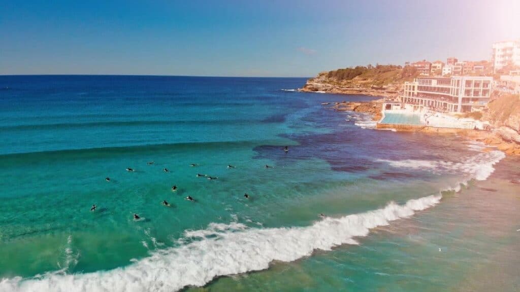 Aerial view of surfers catching waves off the vibrant turquoise coast near a cliff-side urban setting, illustrating the allure of popular surf spots in Australia.