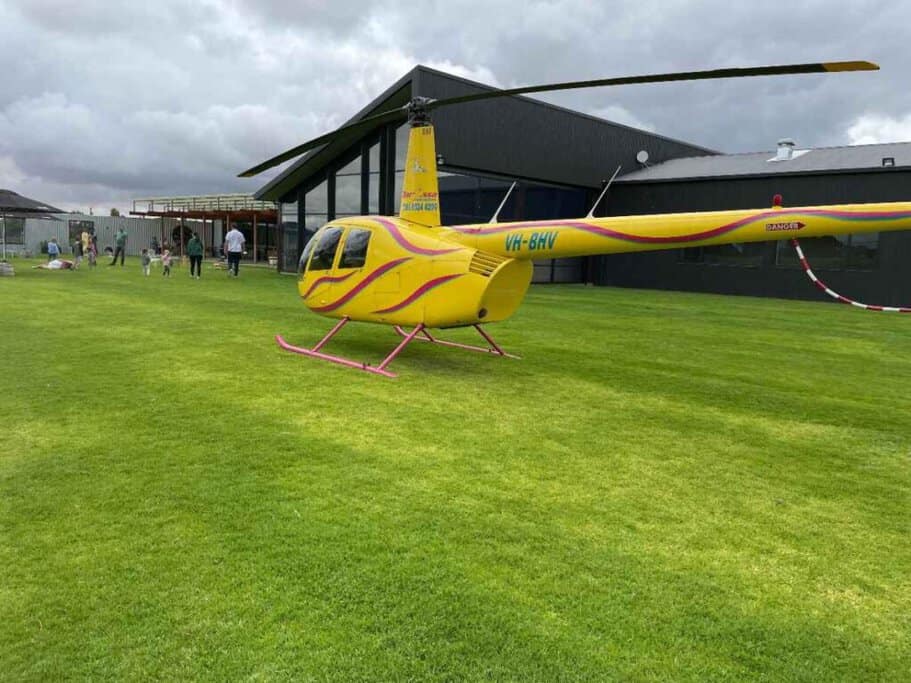 A bright yellow Robinson R44 helicopter with the registration VH-BHV, parked on a lush green lawn at the Artisans of Barossa venue, with people mingling in the background. This scene captures the essence of Barossa Valley helicopter tours, showcasing the unique experience of vineyard visits by air.