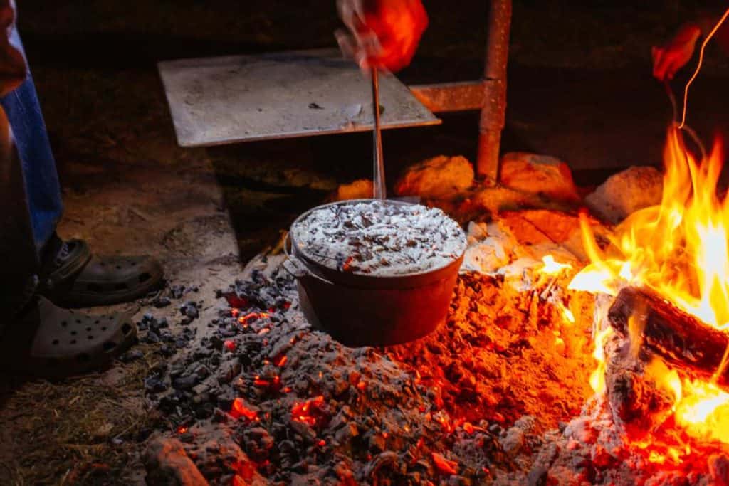 A traditional Australian campfire cooking setup featuring a cast-iron camp oven filled with damper, an iconic Aussie food. The pot is surrounded by hot coals, creating an authentic bush cooking experience. This classic method of baking damper highlights Australia's outdoor culture and history.