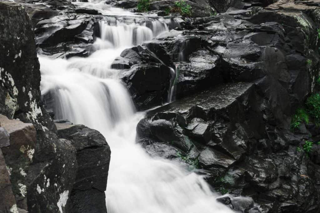 a cascading waterfall flowing over dark, jagged rocks surrounded by patches of greenery. This serene scene is likely a picturesque spot, such as Gardners Falls in Maleny, which highlights why this area is featured in a post about the best waterfalls in Queensland.