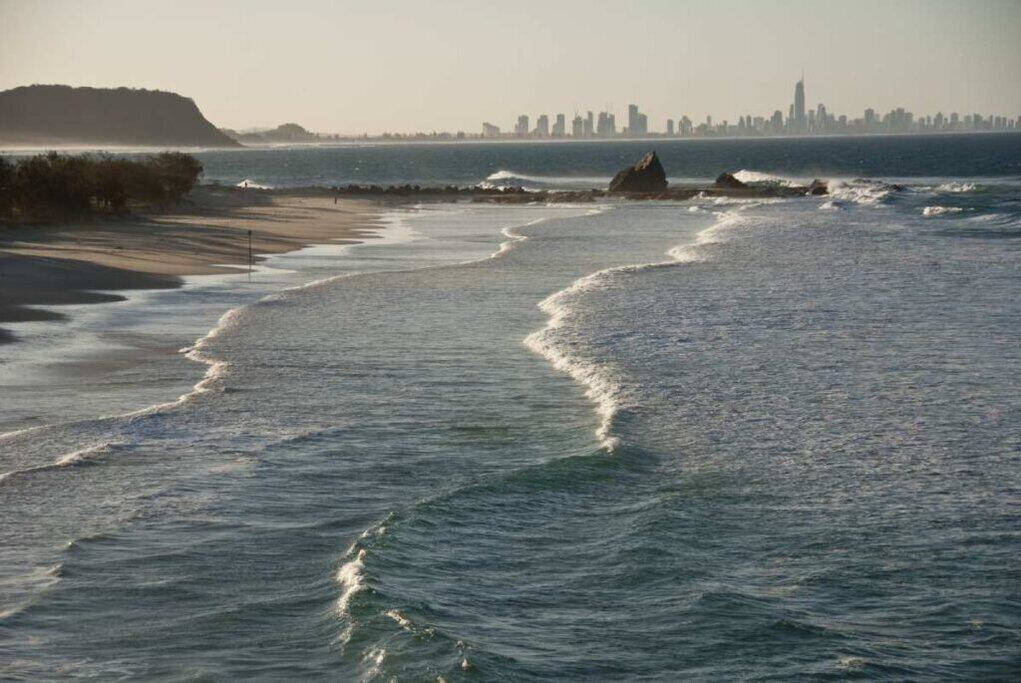 Golden beaches and rolling waves along the Gold Coast, with the skyline of Surfers Paradise visible in the distance under a hazy summer sky. This scene captures the allure of summer in Australia, highlighting the Gold Coast as a prime location for beachgoers and city explorers alike.