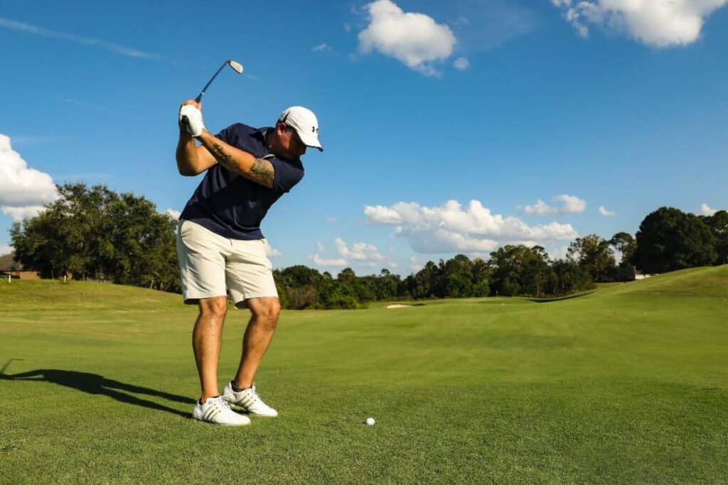 Man in a navy polo shirt and white shorts swinging a golf club on a sunny day at a lush golf course, representing an active day of golfing on a golf and wine tour in Barossa Valley.