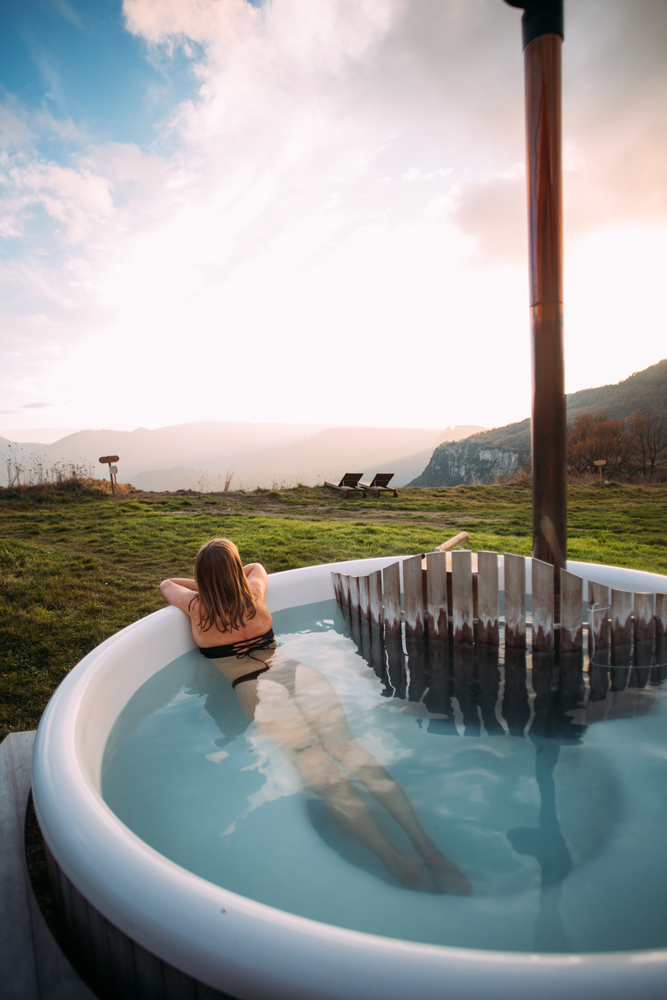 Young beautiful woman in bikini lay inside hipster modern hot tub heated with wood. Amazing retreat location, glamping or spa resort in nature. Outdoors lifestyle, living in countryside. 