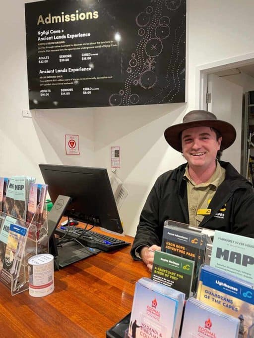 A friendly staff member at the admissions desk of Ngilgi Cave, with pricing information displayed behind him. This image supports a guide to Ngilgi Cave by highlighting the helpful staff and available visitor information.