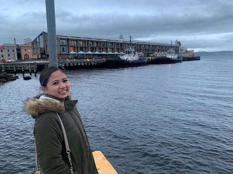 The author, Kate, standing at the Hobart waterfront, with calm waters and cloudy skies, on a day where snow is not present, inviting speculation about the typical climate and whether it snows in Hobart.