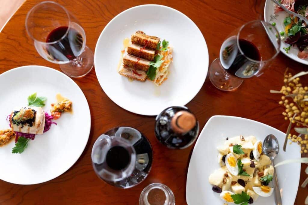 Elegant gourmet dishes paired with fine wines on a wooden table, part of a culinary experience on a Barossa Valley tour, showcasing a diverse spread including seared fish, crispy pork belly, and a creamy potato salad.