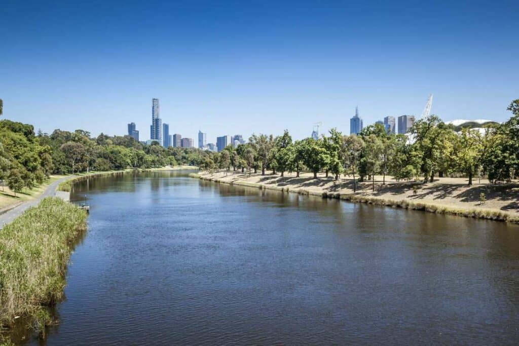 Melbourne's city skyline viewed from across the Yarra River, with lush greenery lining the riverbanks under a bright blue sky. This image represents the vibrant urban and natural landscapes that make Melbourne a top destination to explore during Australia in summer.