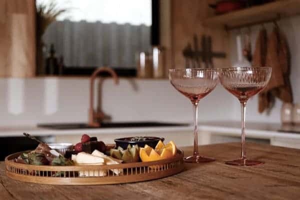 A gourmet platter with a selection of cheeses, fruits, and chocolates, paired with two elegant rose-tinted champagne glasses, set on a wooden kitchen countertop in a luxury cabin near Derby, showcasing the exquisite culinary experiences available to guests.