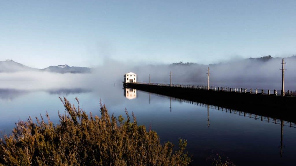 A misty morning view of Pumphouse Point on Lake St Clair, with the white pumphouse building reflecting on the still water and surrounded by low-lying fog and mountains. This serene and dramatic landscape highlights one of the most Instagrammable spots in Tasmania, perfect for moody, reflective photography.